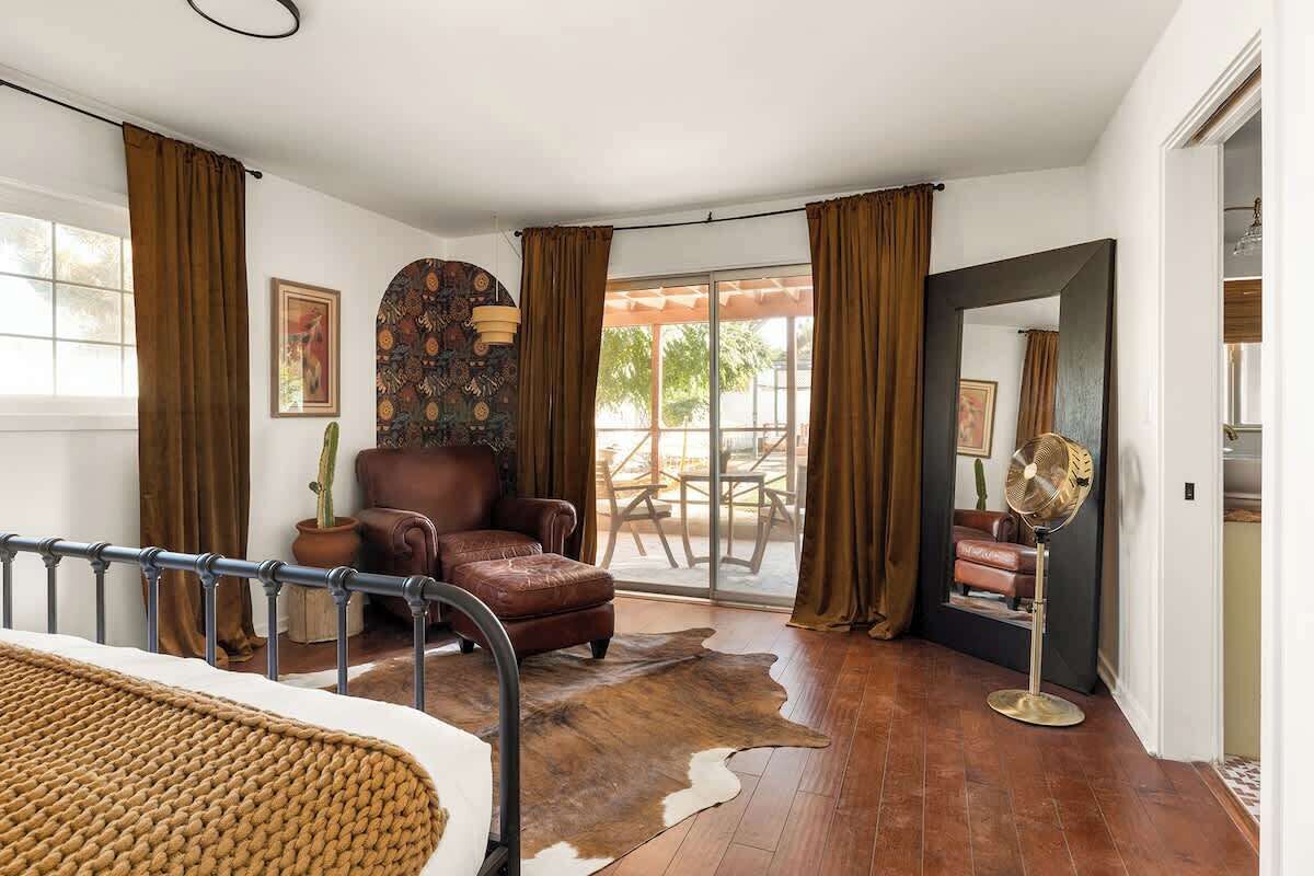 A room with a bed, a leather armchair, a cowhide rug, a fan, and a view of the outdoors through a glass door.