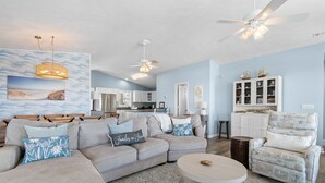 A spacious living room with a large sectional sofa, a round coffee table, and a patterned rug. There is a dining area with a wooden table and chairs, and a kitchen in the background.