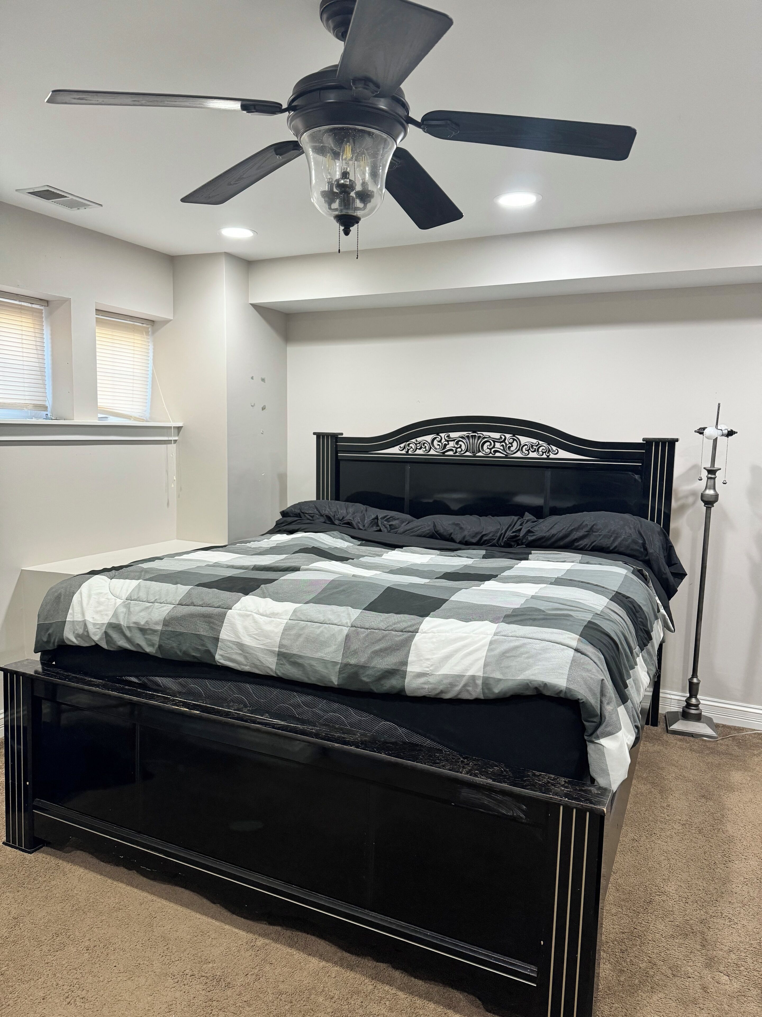Une chambre à coucher avec un cadre de lit noir, une couvre-lit à carreaux, un ventilateur de plafond et une lampe sur pied.