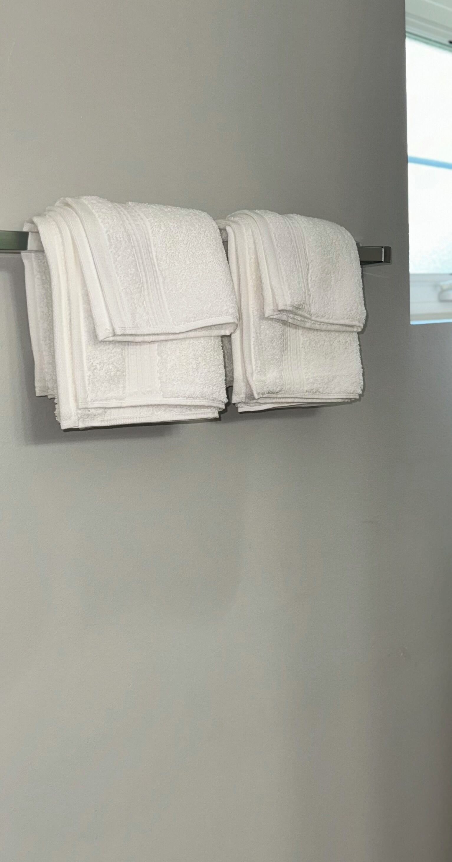 Deux serviettes blanches, pliées avec soin, sont suspendues à un porte-serviettes en métal, le tout adossé à un mur uni.