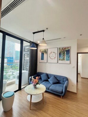 A modern living room with a blue sofa, a round coffee table with a vase of flowers, and a large window offering a city view. - Asahi Luxstay Vinhomes Skylake APT 2br (Hanoi)