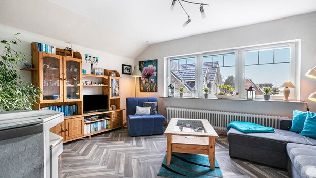 Un salon moderne avec un parquet en chevrons, un canapé, une table basse, une bibliothèque et une grande fenêtre donnant sur des immeubles.