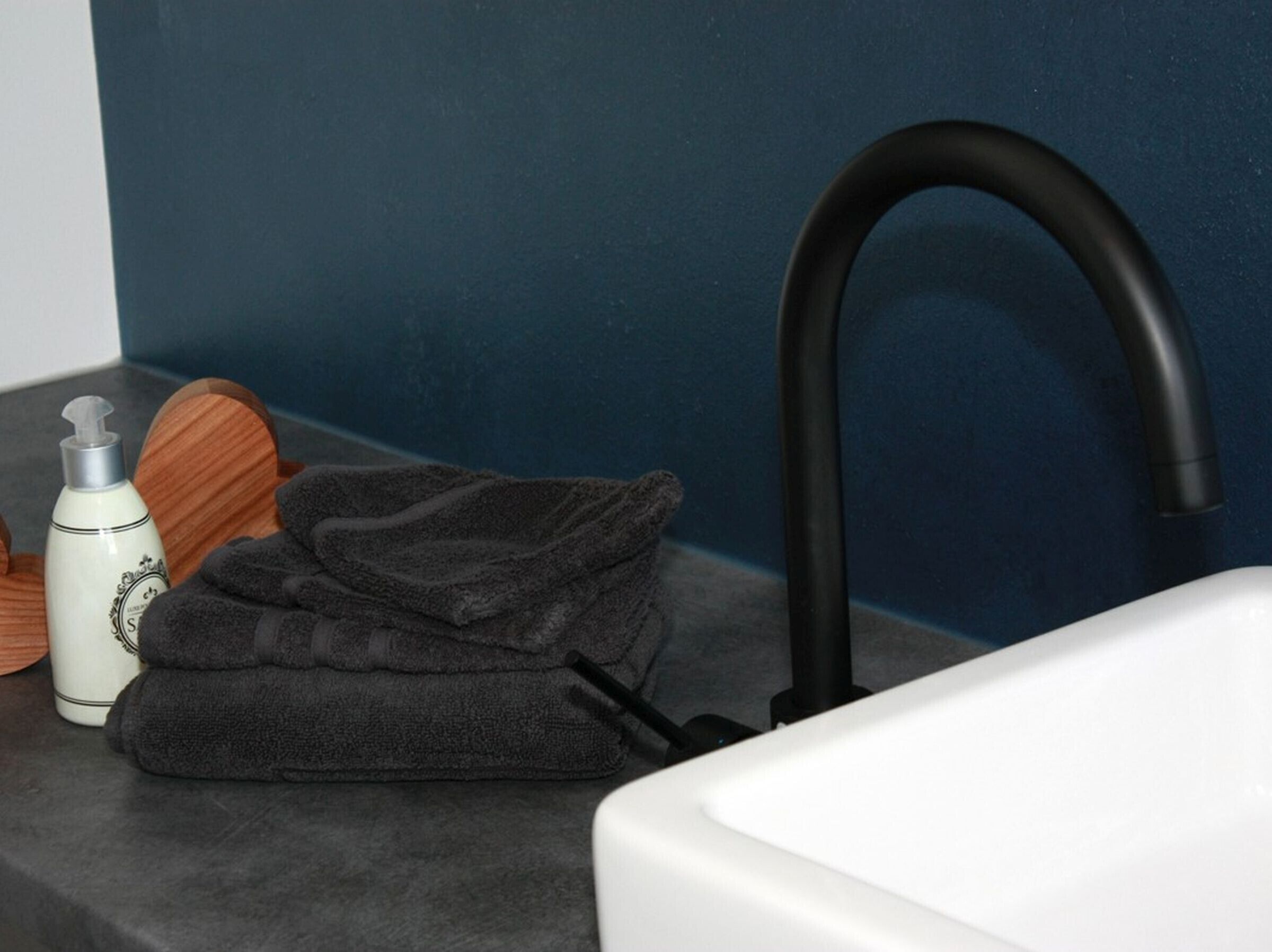 Une salle de bain avec un robinet noir, un lavabo blanc, une serviette grise et un flacon de lotion posé sur un plateau en bois.