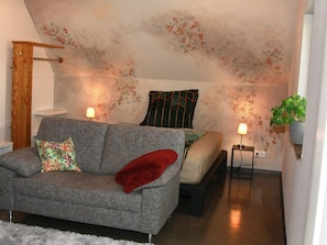A room with a sofa, a bed, a small table, a hanging plant, and a unique ceiling design.