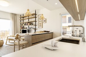 A modern kitchen with a central island, wooden shelving, pendant lighting, and a view to the outside.