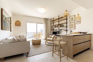 A modern living room with a sofa, chairs, a dining area, and a kitchen with wooden shelves and pendant lights.