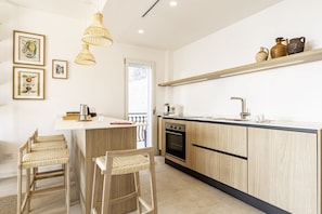 A modern kitchen with wooden cabinets, a built-in oven, and a black countertop. There are two wicker chairs and a hanging light fixture.