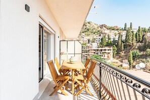 A balcony with a wooden table and chairs, a view of a hillside town, and a door leading to an interior space.