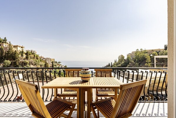 A wooden table and chairs on a balcony with a view of the sea and hillside buildings.