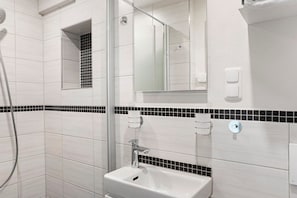 A bathroom with white tiles, a glass shower enclosure, a sink with a mirror above it, and a black and white tiled accent strip.