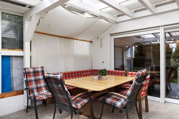 A covered patio with a wooden table and plaid cushioned chairs, a potted plant, and a view through a glass door.