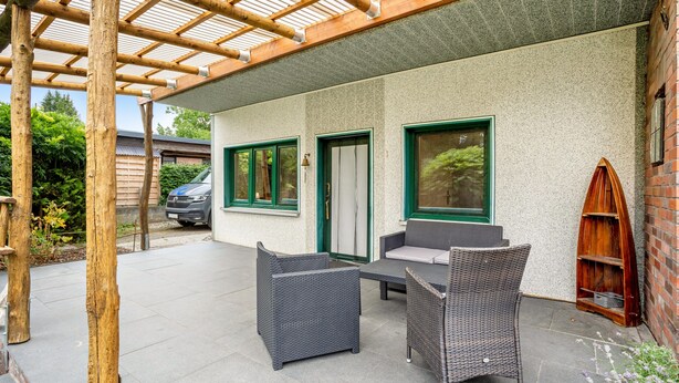 Un patio couvert avec deux fauteuils en osier et une table, une voiture garée à l’extérieur et un bâtiment doté d’un mur texturé et de fenêtres à cadre vert.