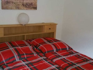 A bed with a red plaid bedspread, a bedside table with a lamp, and a wall-mounted artwork.