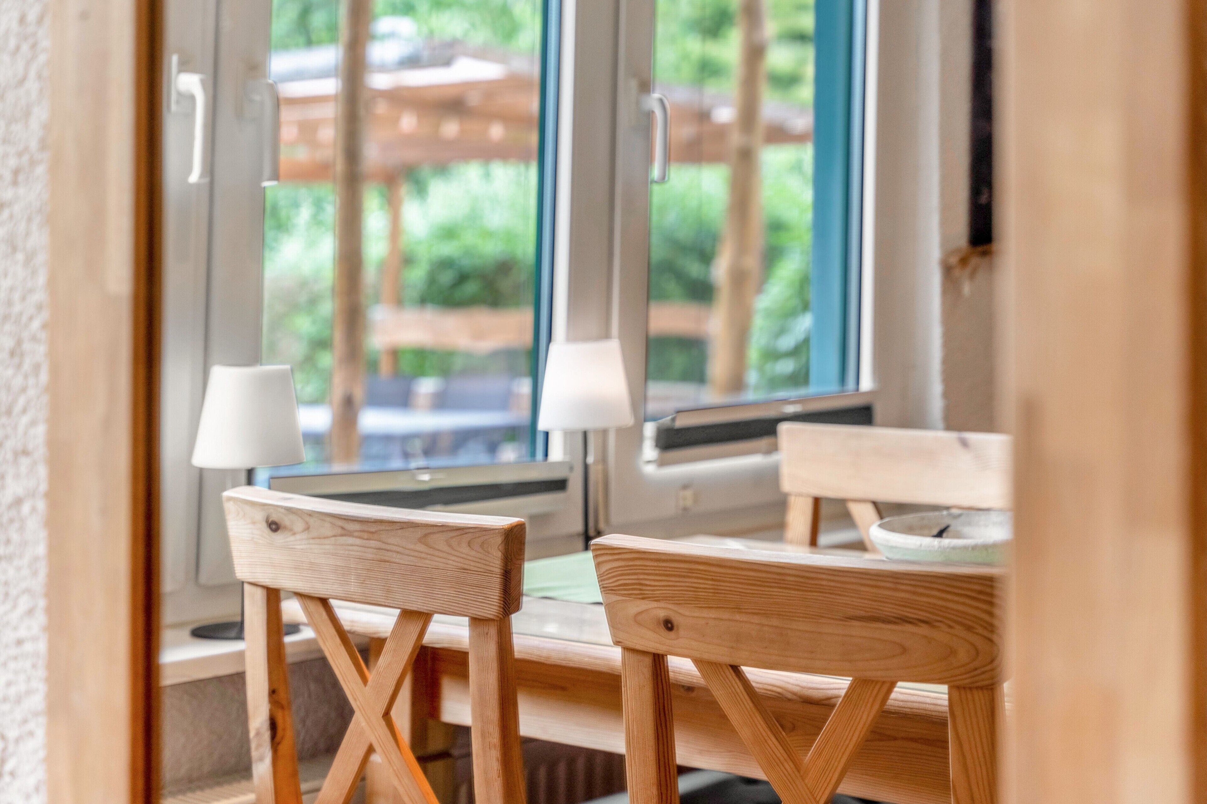 A wooden table with two chairs, a window with a view of greenery, and a white lamp on the table.