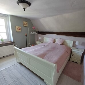 Ein Schlafzimmer mit Bett, Nachttischen, Heizkörper, Fenster und einer Deckenleuchte.