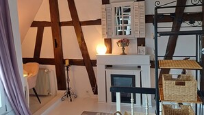 A room with exposed wooden beams, a fireplace, a white mantelpiece, and a metal shelf with baskets. - Apartment 'Weinbergsblick' with Private Terrace, Private Garden and Balcony (Geisenheim)