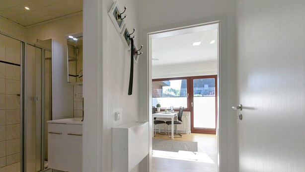 A modern bathroom with a shower, a toilet, and a sink. There is a hallway leading to a dining area with a table and chairs.