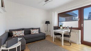 A modern living room with a grey sofa, a small white table, a dining table with a chair, a floor lamp, and a large window.