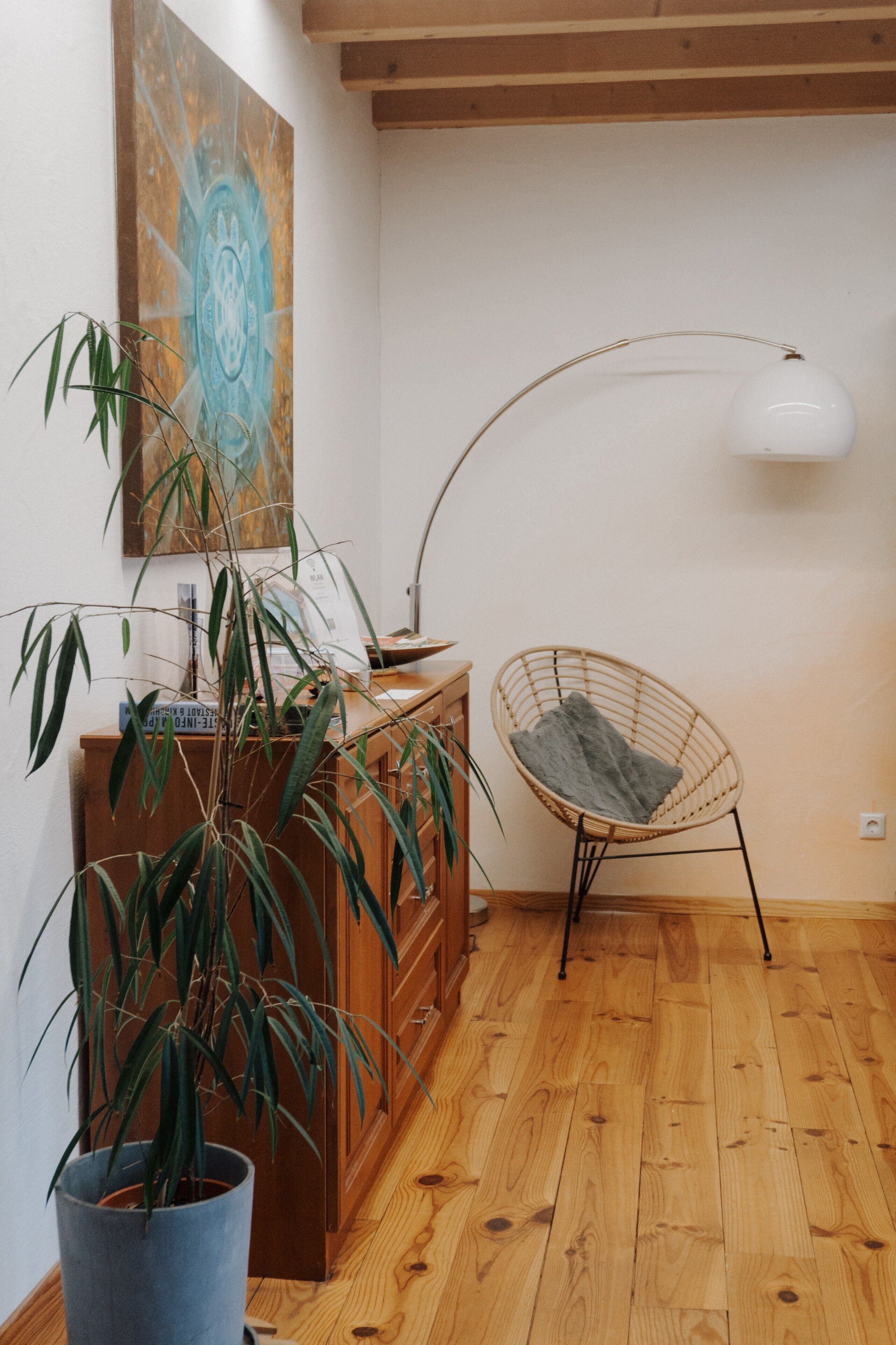 Een kamer met houten vloeren, een bureau met lades, een rieten stoel, een kamerplant en een wandlamp.