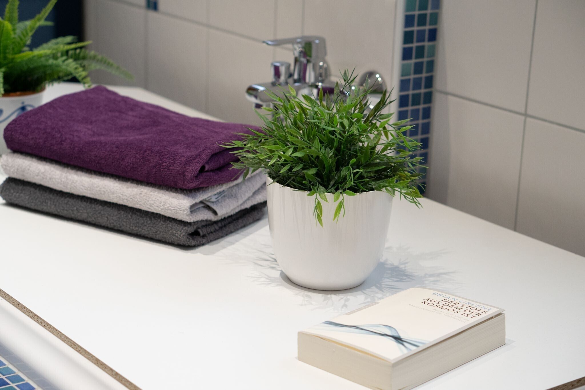 A bathroom with a white sink, a purple towel, a grey towel, a white book, and a green plant in a white pot.