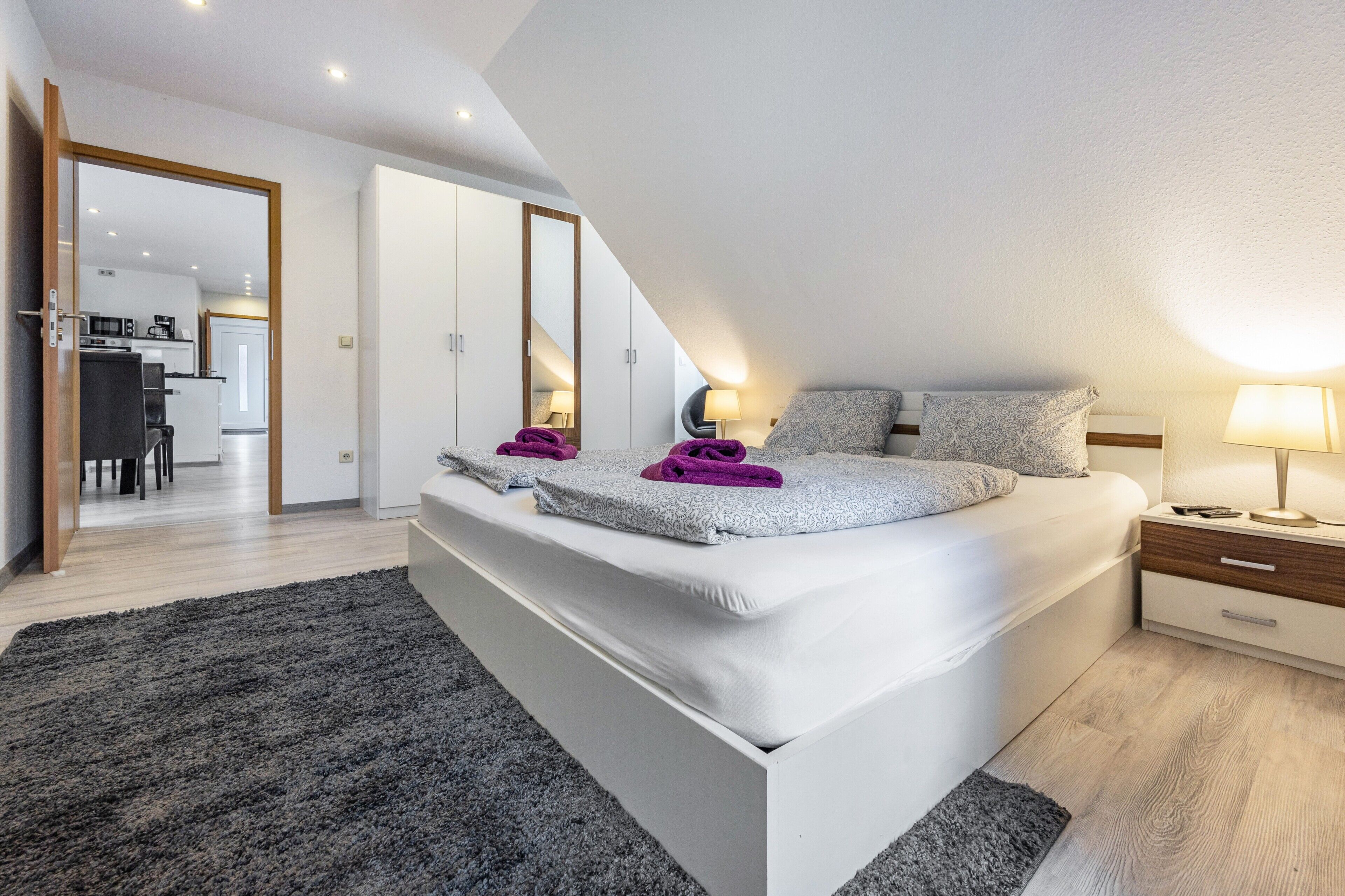 A bedroom with a large bed, a nightstand, a lamp, and a view of the kitchen and dining area.
