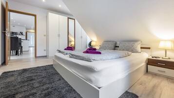 A bedroom with a large bed, a nightstand, a lamp, and a view of the kitchen and dining area.