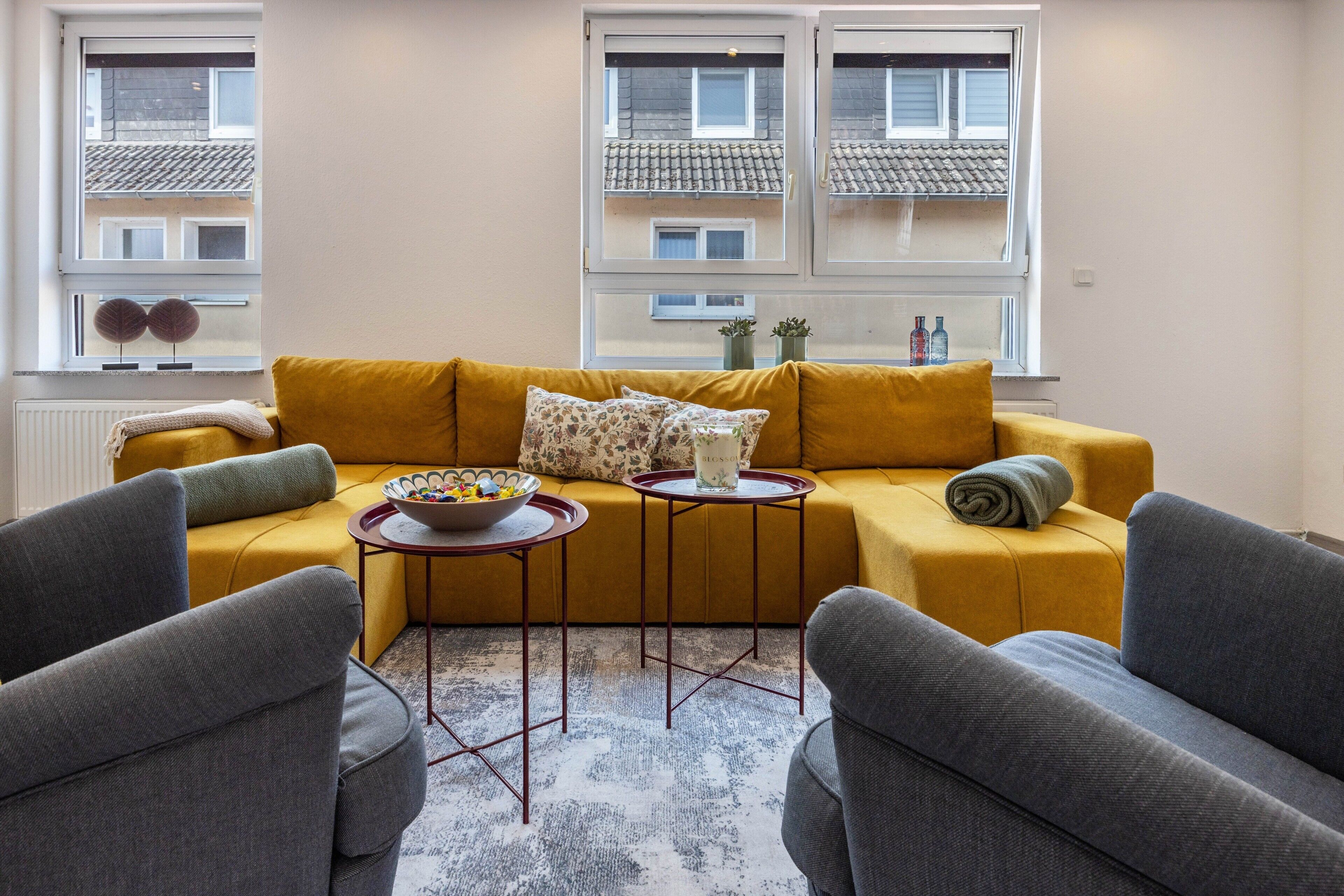A living room with a mustard sofa, two armchairs, a coffee table, and a view of a building through windows.
