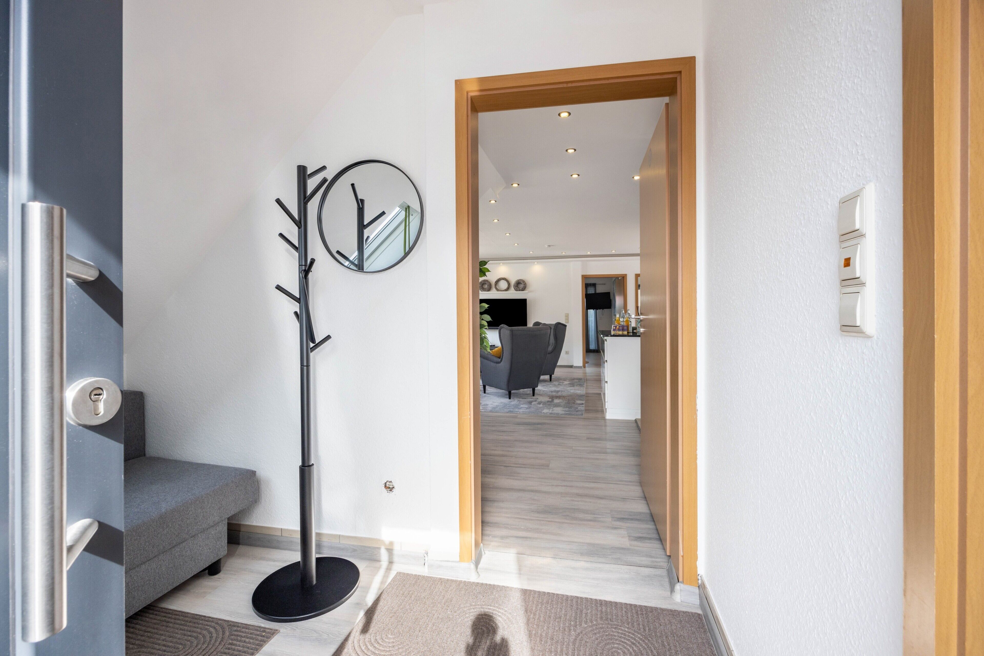 Ein modernes Interieur mit einem grauen Sofa, einem Kleiderständer und einer Holztür, die in einen Wohnbereich führt.