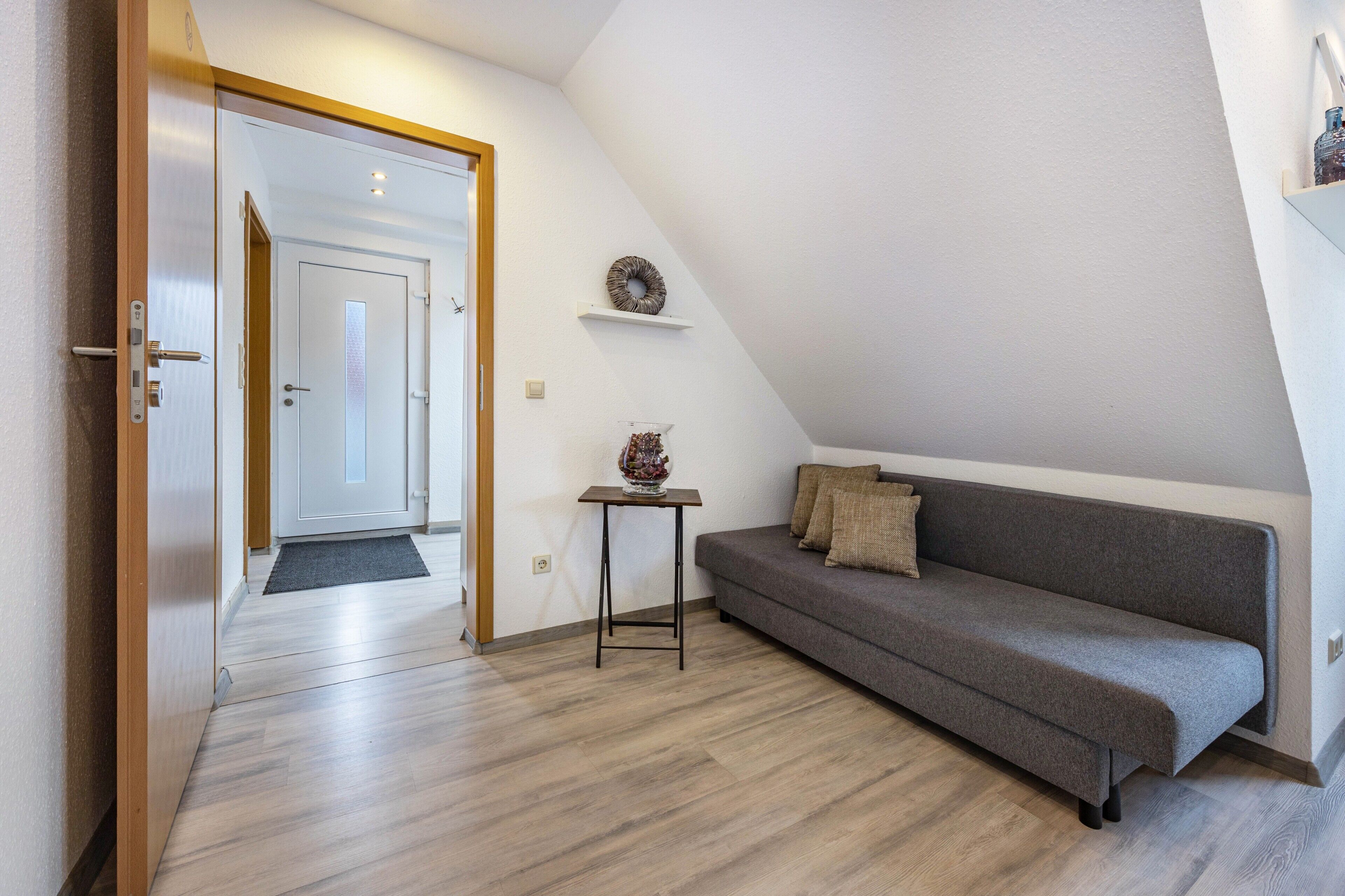 Una habitación moderna con suelo de madera, un sofá gris, una pequeña mesa con un cuenco decorativo y una puerta que da a otra habitación.
