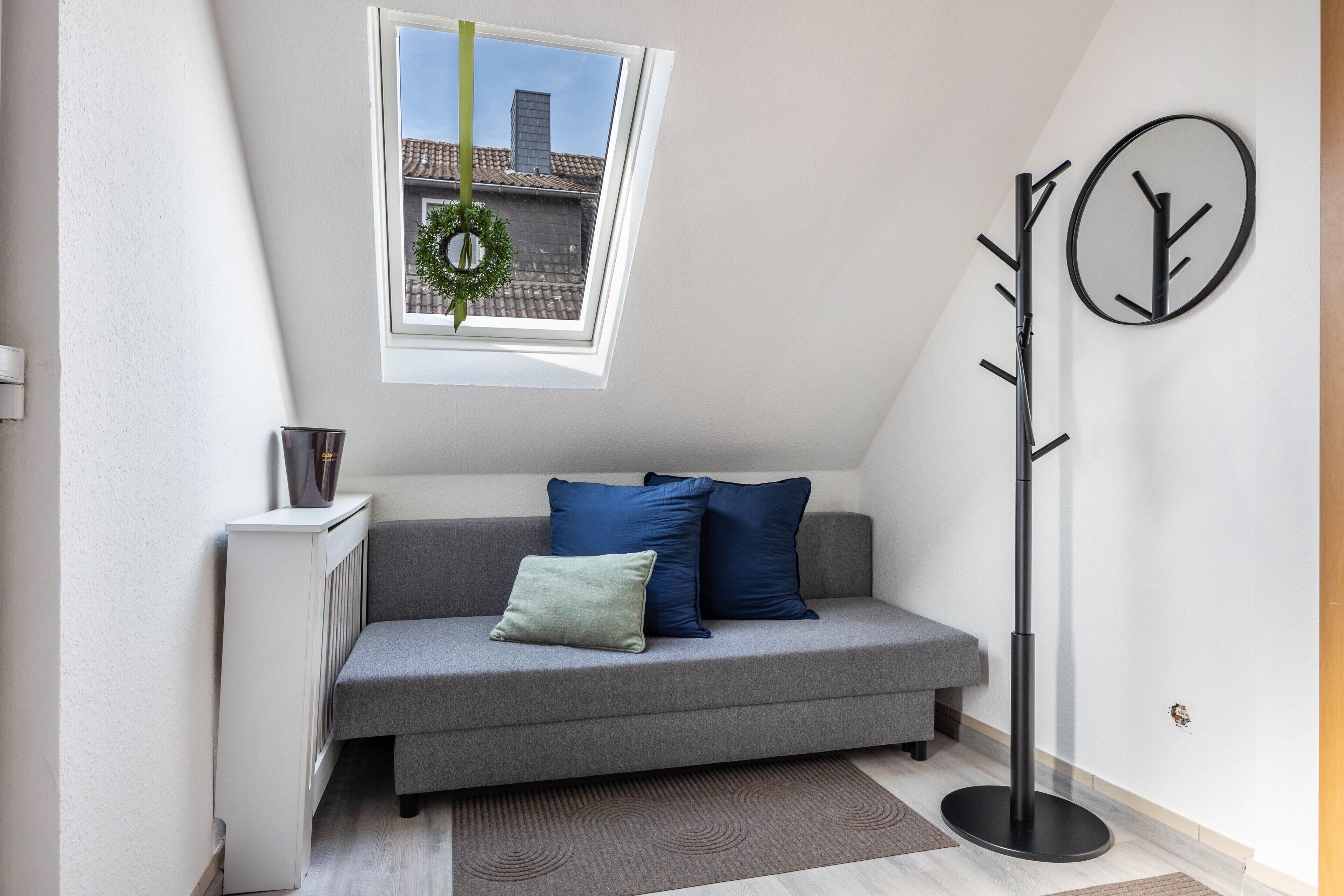 Un salon moderne avec un canapé gris, un petit radiateur, une fenêtre décorée d’une guirlande et un porte-manteau.