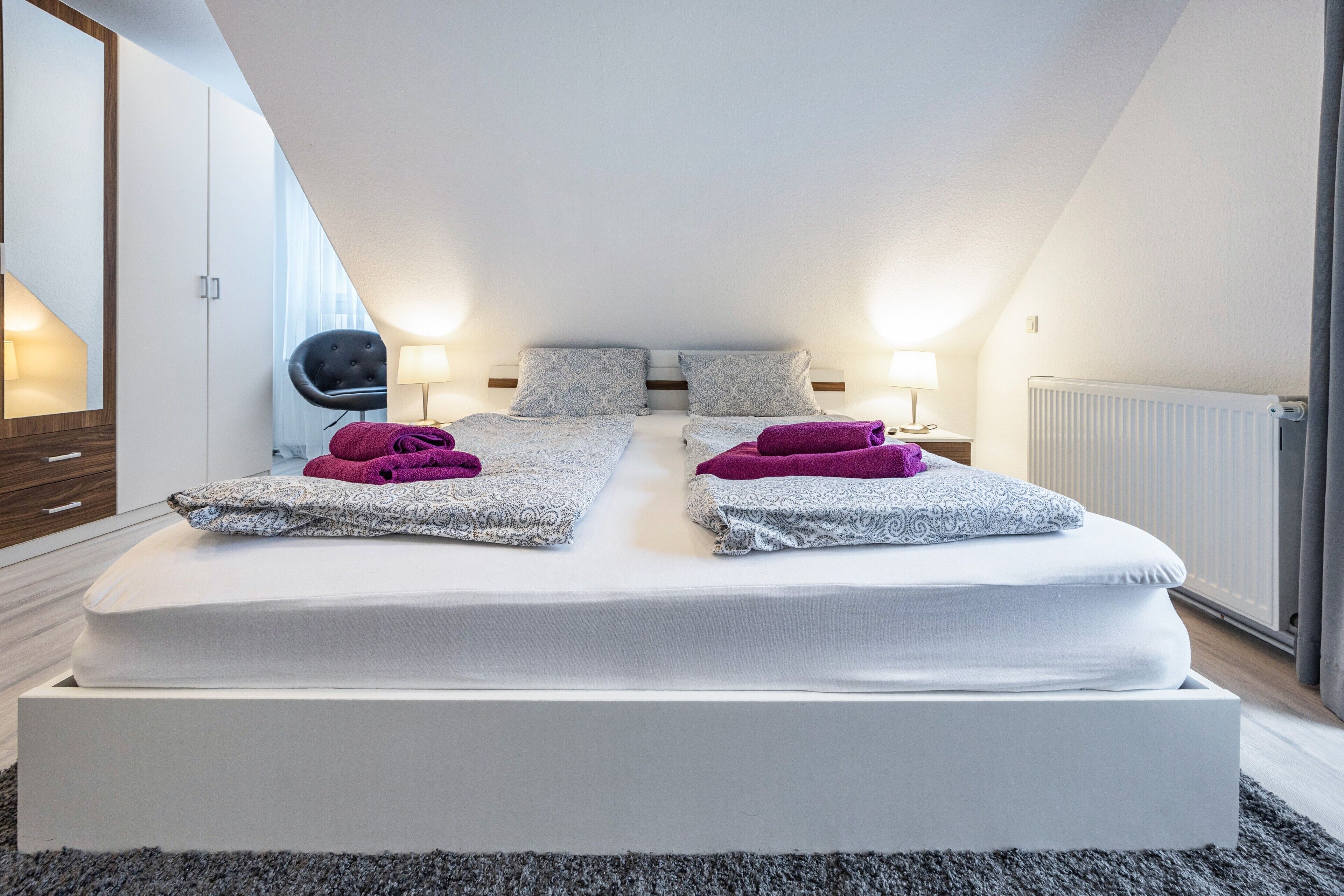 A modern bedroom with a large bed, two bedside tables with lamps, a chair, and a radiator.
