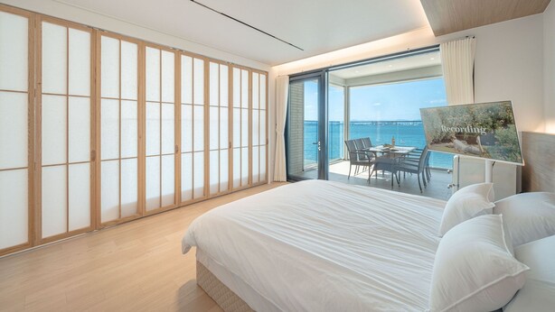 Una camera da letto moderna con un letto grande, una porta scorrevole in vetro che dà su un balcone con vista sul mare e un quadro appeso alla parete.