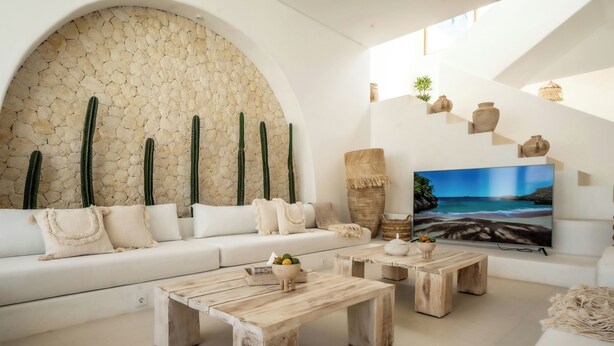 Un soggiorno moderno con un divano bianco, tavolini in legno, una parete in pietra decorata con cactus, un televisore e una scala abbellita con ceramiche.