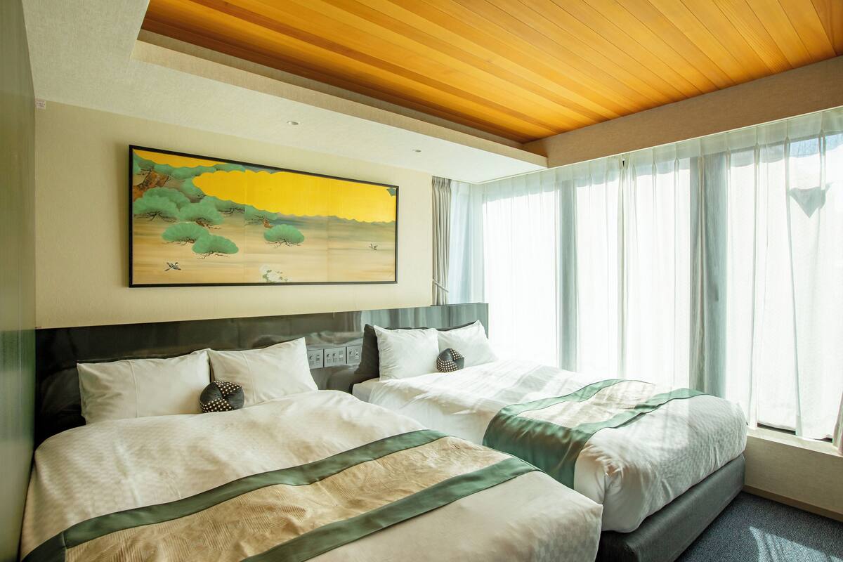 Camera d'albergo con due letti, un quadro appeso al muro e un'ampia finestra con tende.