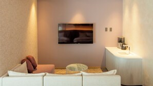 Een moderne kamer met een flatscreen televisie aan de muur, een witte bank met kussens, een klein rond tafeltje en een witte console met een wastafel.