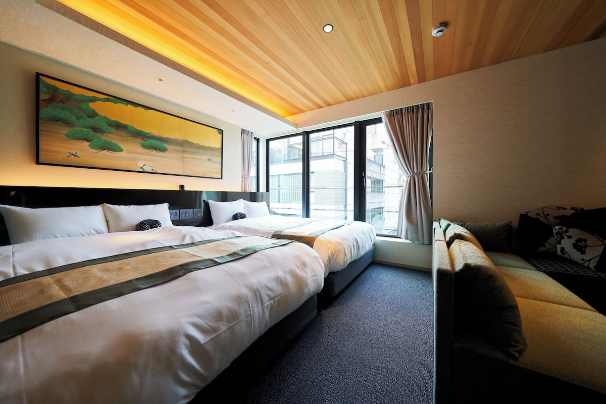 Habitación de hotel con dos camas, un sofá, un ventanal grande y un cuadro en la pared.