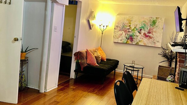 En hyggelig stue med trægulv, en sort sofa, et spisebord med stole og et billede på væggen.