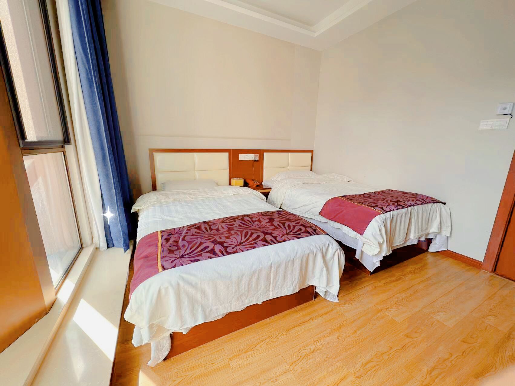 A hotel room with two beds, wooden flooring, a window with curtains, and a white wall.