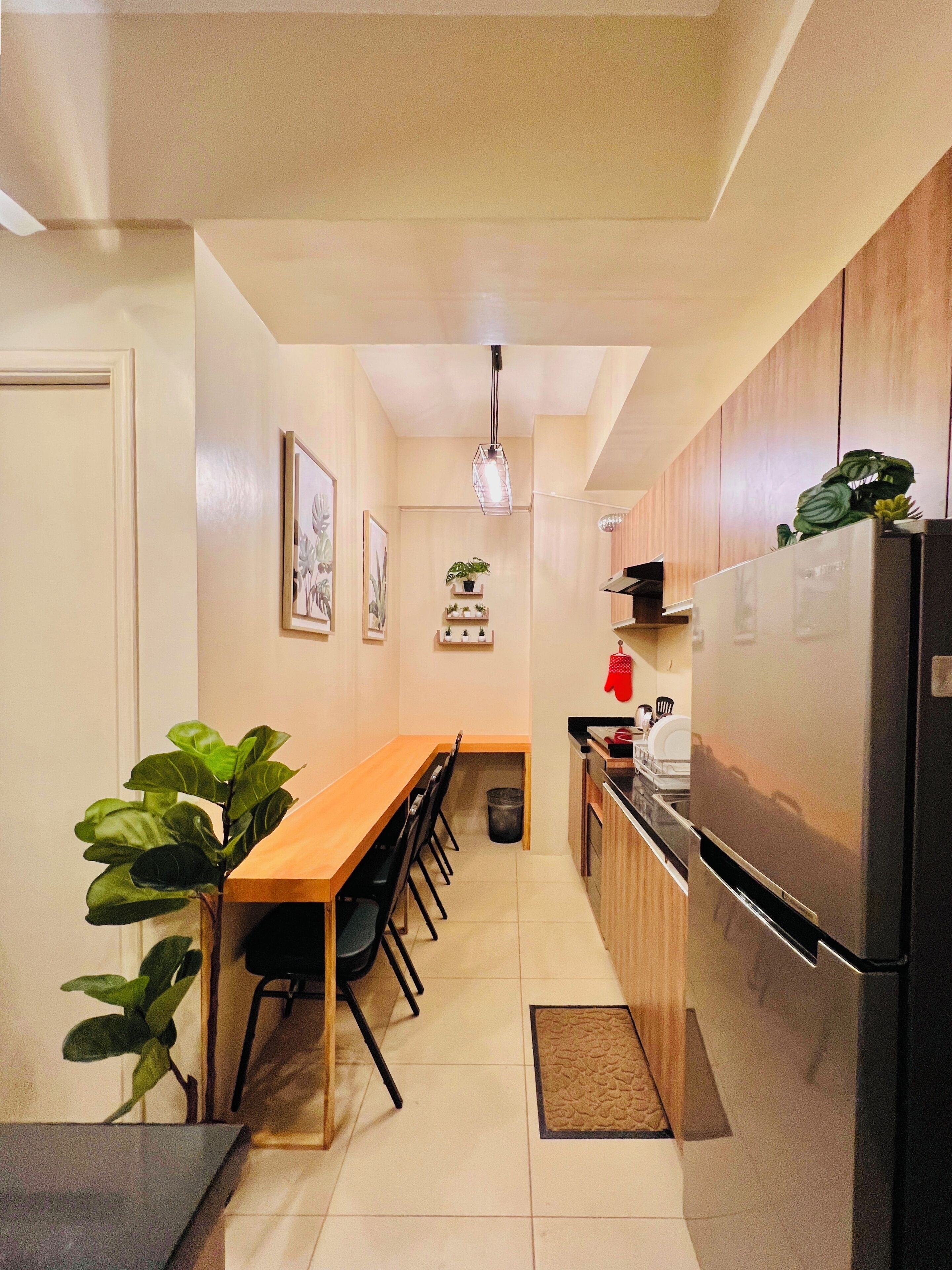 Een kleine keuken met een eethoek, met een lange houten tafel, stoelen, een koelkast en een hanglamp.