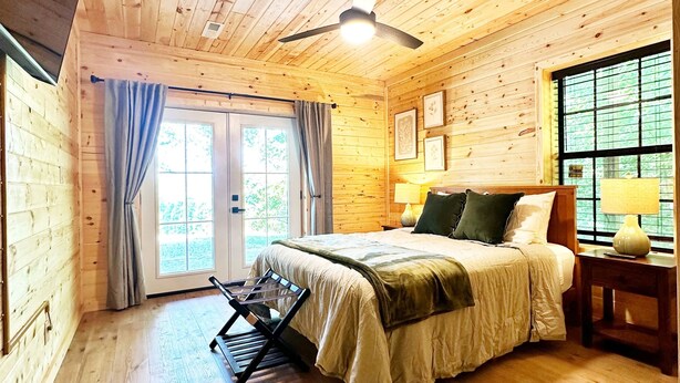 Une chambre dans une cabane en bois, avec un lit, un ventilateur de plafond, une fenêtre avec des rideaux et un plancher en bois.