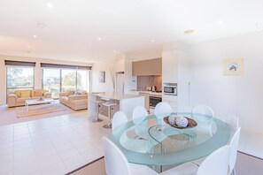 A modern living room with a glass dining table, white sofas, and a kitchen in the background. - 3 Bedroom Townhouse - Pool Access (Torquay)