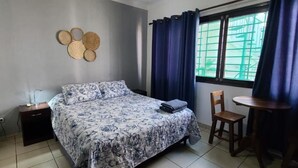 Un dormitorio con cama, mesita de noche, silla, una pequeña mesa y ventana con cortinas.