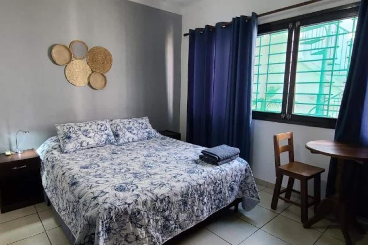 A bedroom with a bed, a bedside table, a chair, a small table, and a window with curtains.
