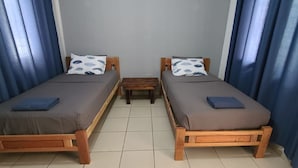 Dos camas de madera con colchones grises y almohadas azules, una mesita de madera y cortinas azules.