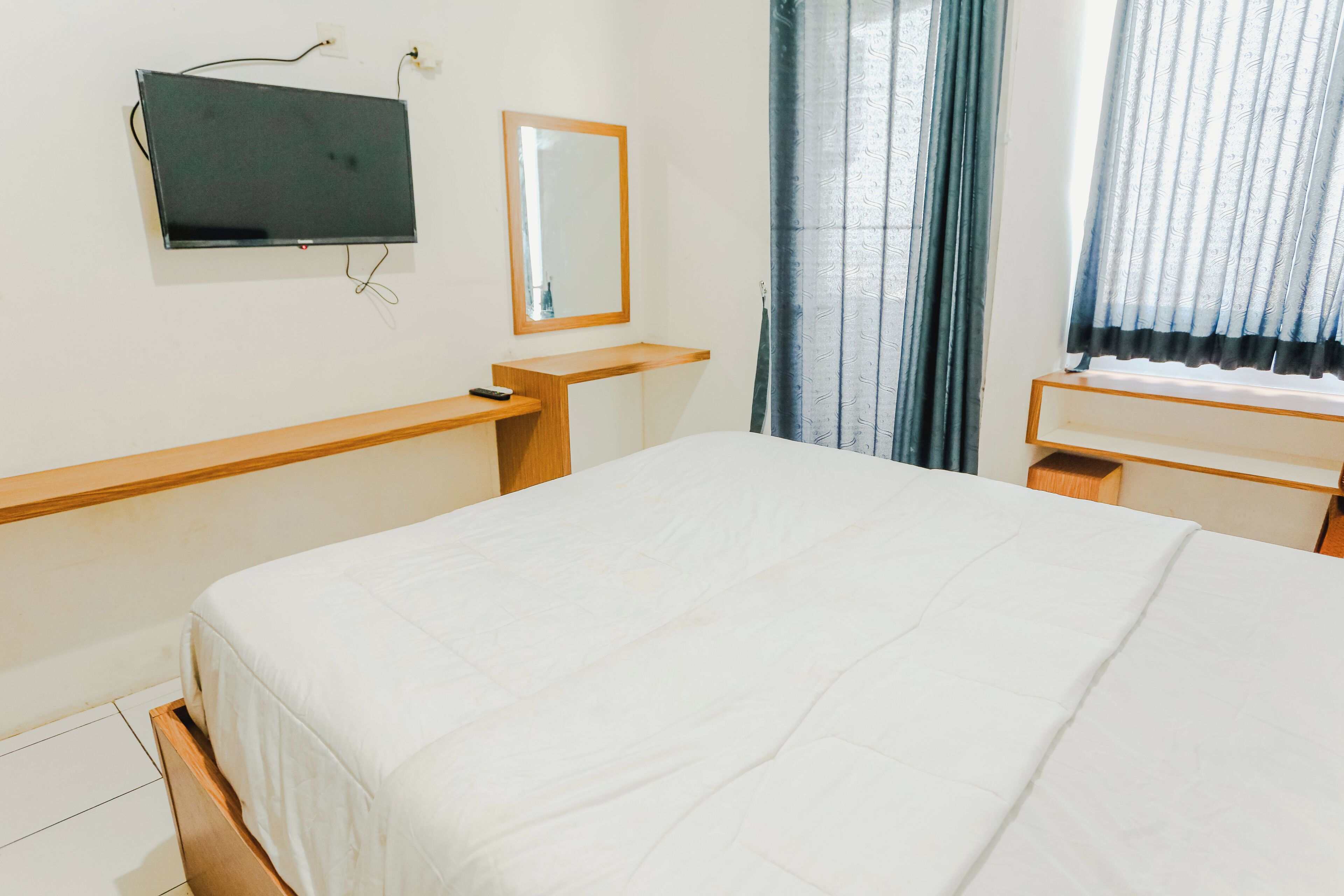 A hotel room with a bed, a TV mounted on the wall, a mirror, and a window with curtains.