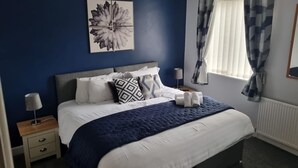 Une chambre à coucher comprenant un lit, une table de chevet, une lampe, une fenêtre avec des rideaux et une œuvre d’art murale.