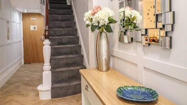 Un couloir moderne avec un escalier en bois, un vase rempli de fleurs et un mur décoratif composé de miroirs.