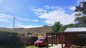 Un coche rojo estacionado frente a una cabaña de madera con techo metálico, rodeada por una valla de madera y un césped.
