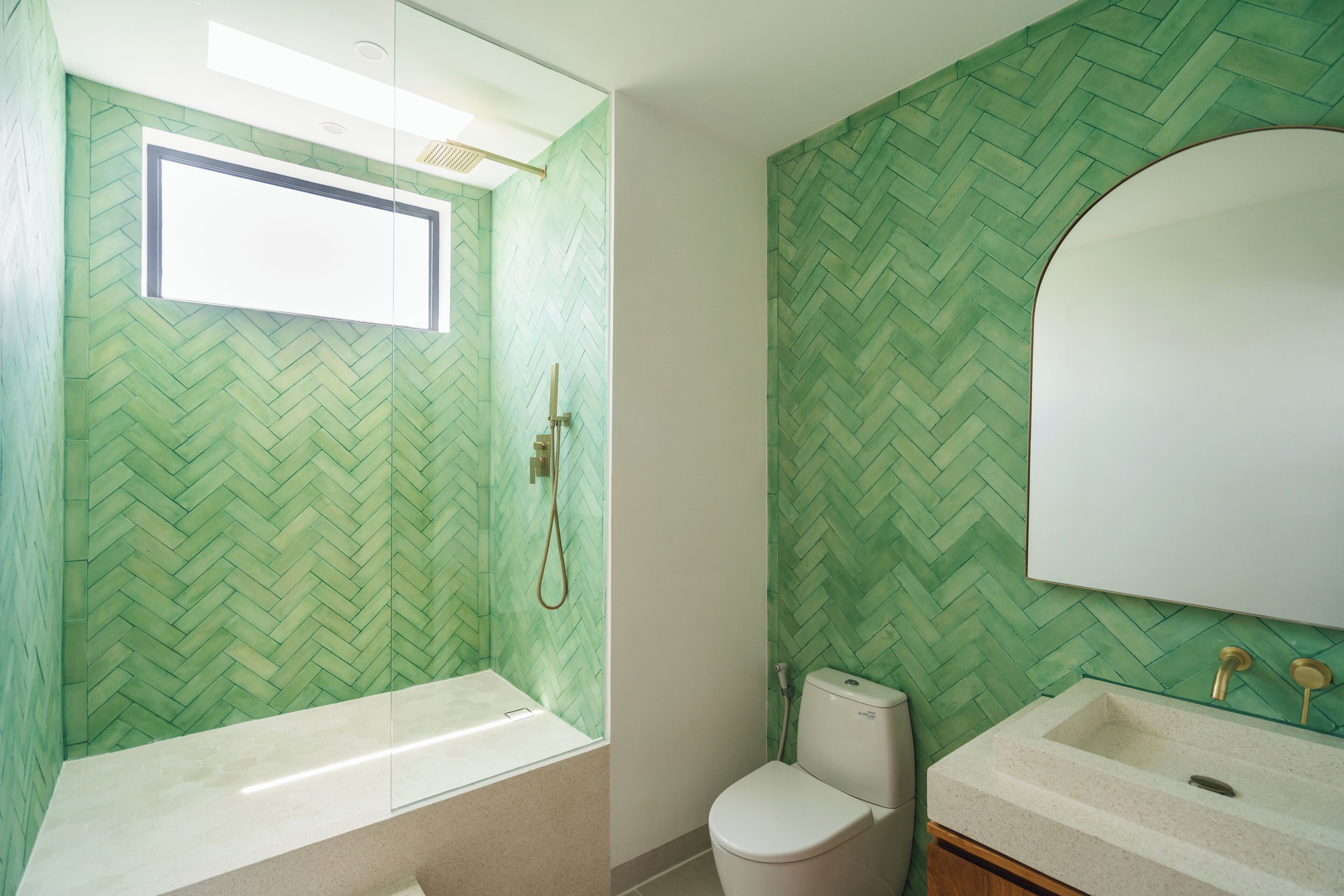 A bathroom with a herringbone-patterned green wall, a window above the bathtub, a toilet, and a sink with a mirror.