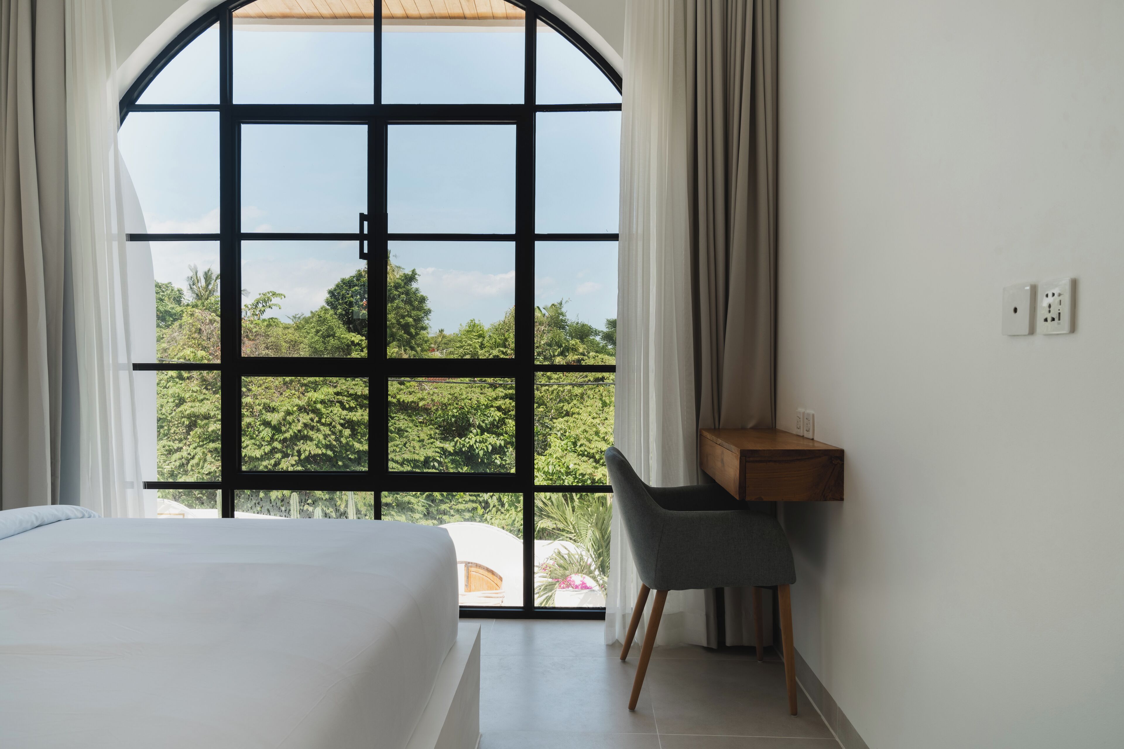 A bedroom with a large arched window, a bed, a desk, and a chair.